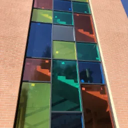 Wysoki, wąski okno z kolorowymi szybami w odcieniach czerwieni, zieleni, niebieskiego i żółtego, wkomponowane w ceglaną ścianę budynku, z widocznymi refleksami światła i nieba.