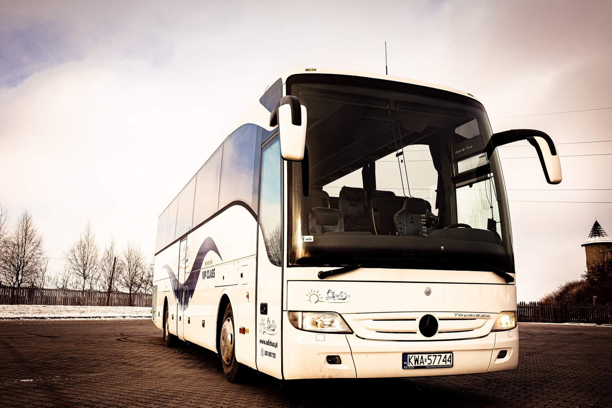 Biały autokar turystyczny Mercedes-Benz Tourismo z napisem VIP Class i logo firmy na tle kostki brukowej i zimowego krajobrazu z wieżą w oddali.