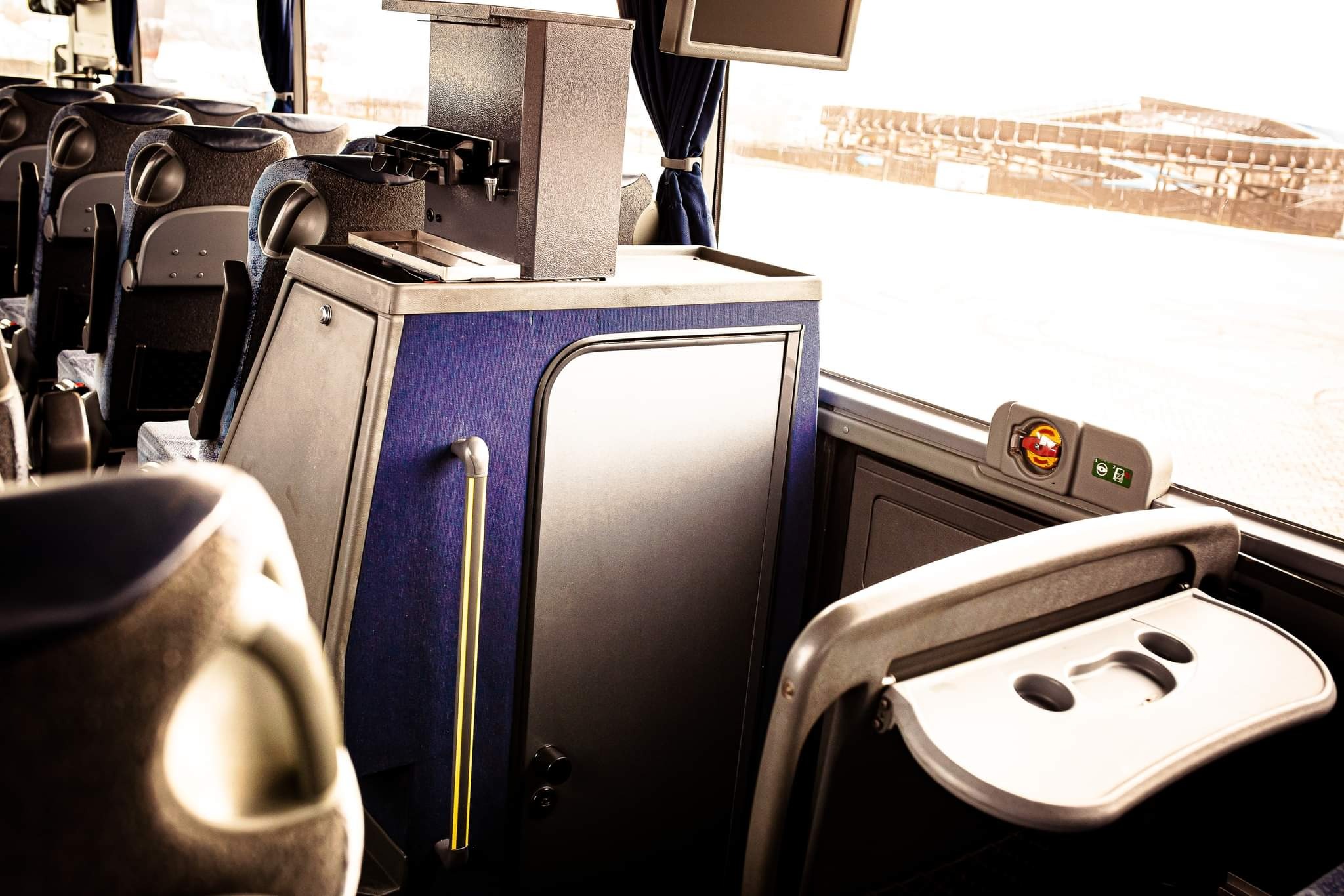 Wnętrze autobusu dalekobieżnego z widocznymi fotelami, stolikiem, zasłoną w oknie i automatem do napojów.