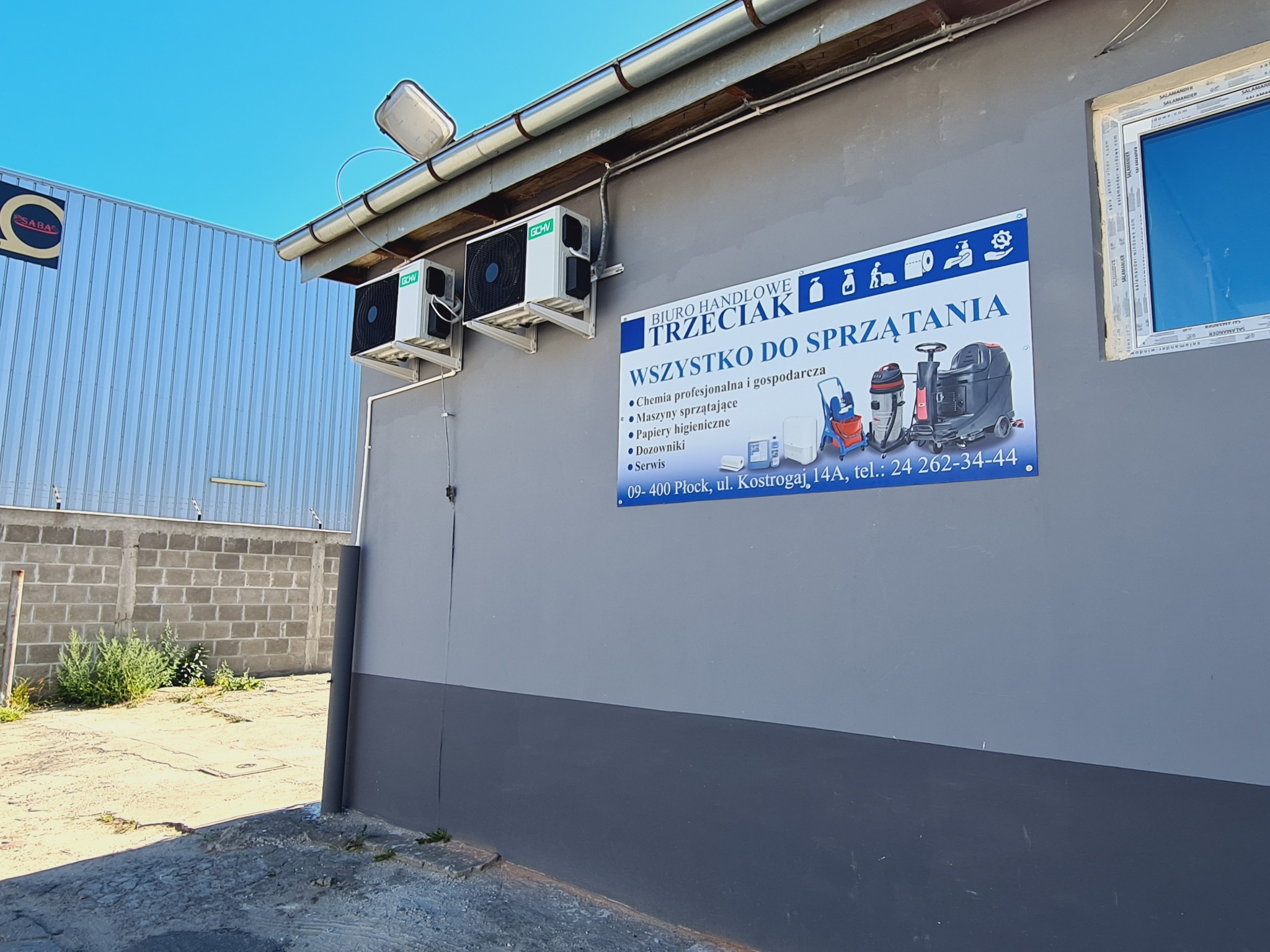 Zewnętrzny widok budynku firmy oferującej artykuły do sprzątania, z widocznym szyldem informacyjnym prezentującym ofertę i danymi kontaktowymi, oraz dwoma klimatyzatorami na ścianie.