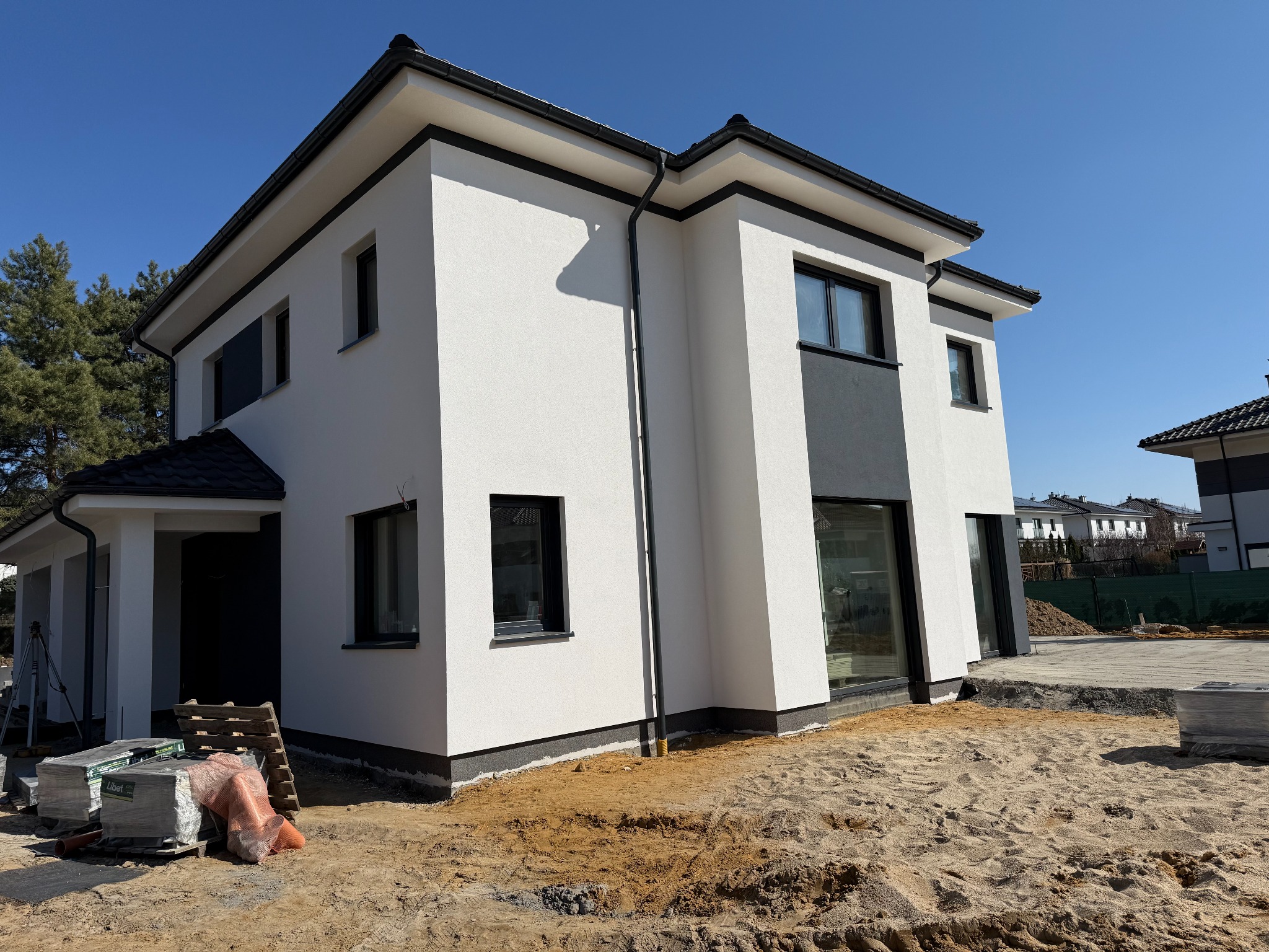 Biały dom z czarnymi akcentami elewacyjnymi w trakcie prac wykończeniowych. Widoczne materiały budowlane i przygotowany teren wokół budynku.