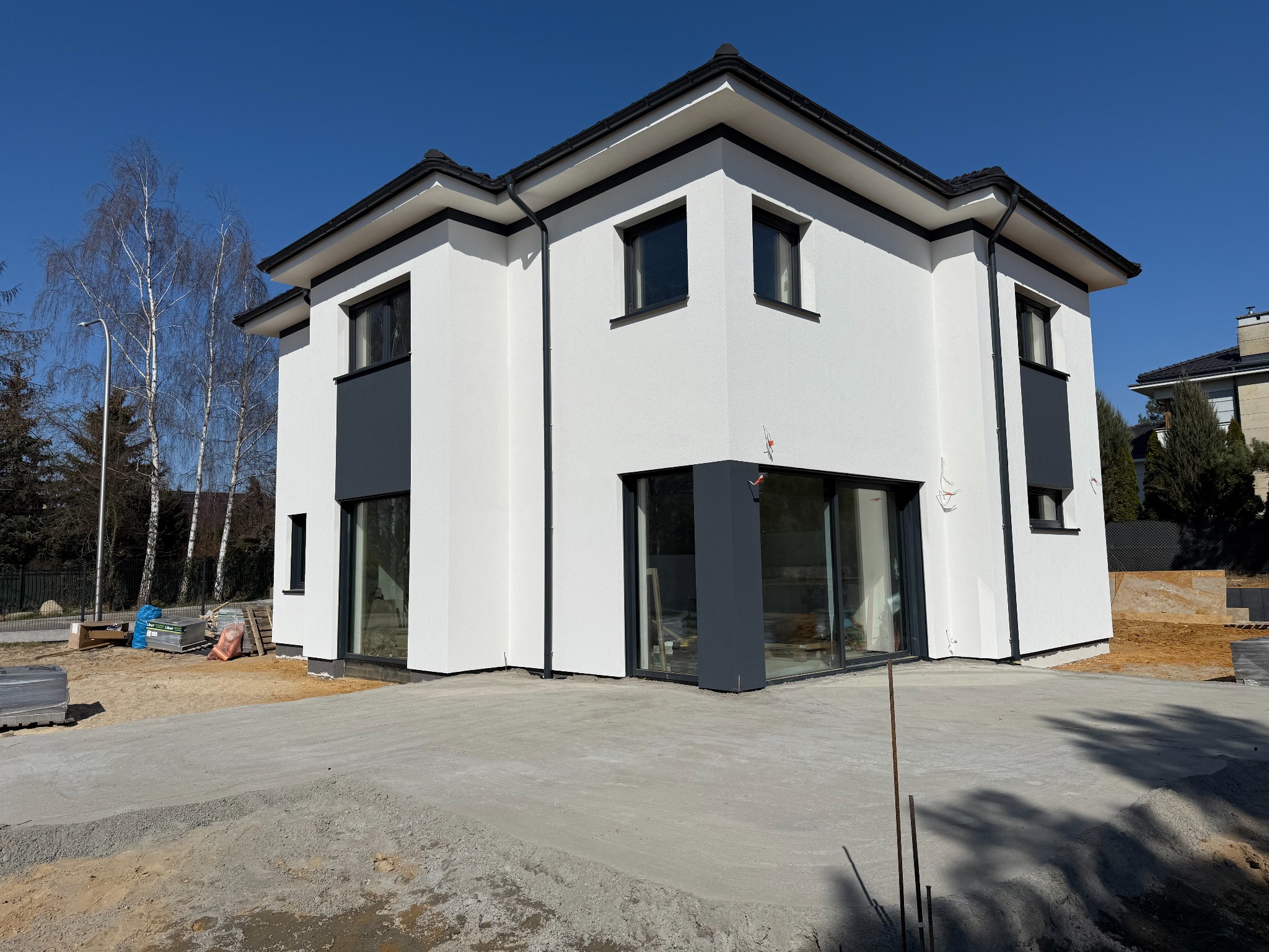 Dwukondygnacyjny dom z białą elewacją i antracytowymi akcentami. Widoczne duże okna i świeżo wylany betonowy podjazd. W tle drzewa i błękitne niebo.