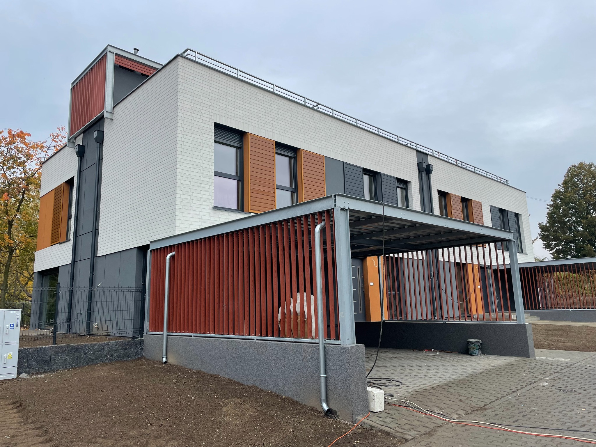 Nowoczesny budynek z elewacją z białej cegły i drewnianymi żaluzjami okiennymi. Widoczny fragment podjazdu z kostki brukowej i elementy wykończeniowe elewacji.