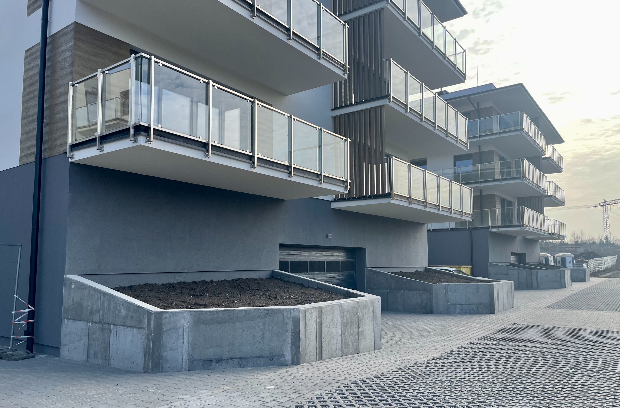 Nowoczesny budynek wielorodzinny z balkonami o szklanych balustradach i szarą elewacją. Widoczne betonowe donice z ziemią i kostka brukowa na podjeździe.