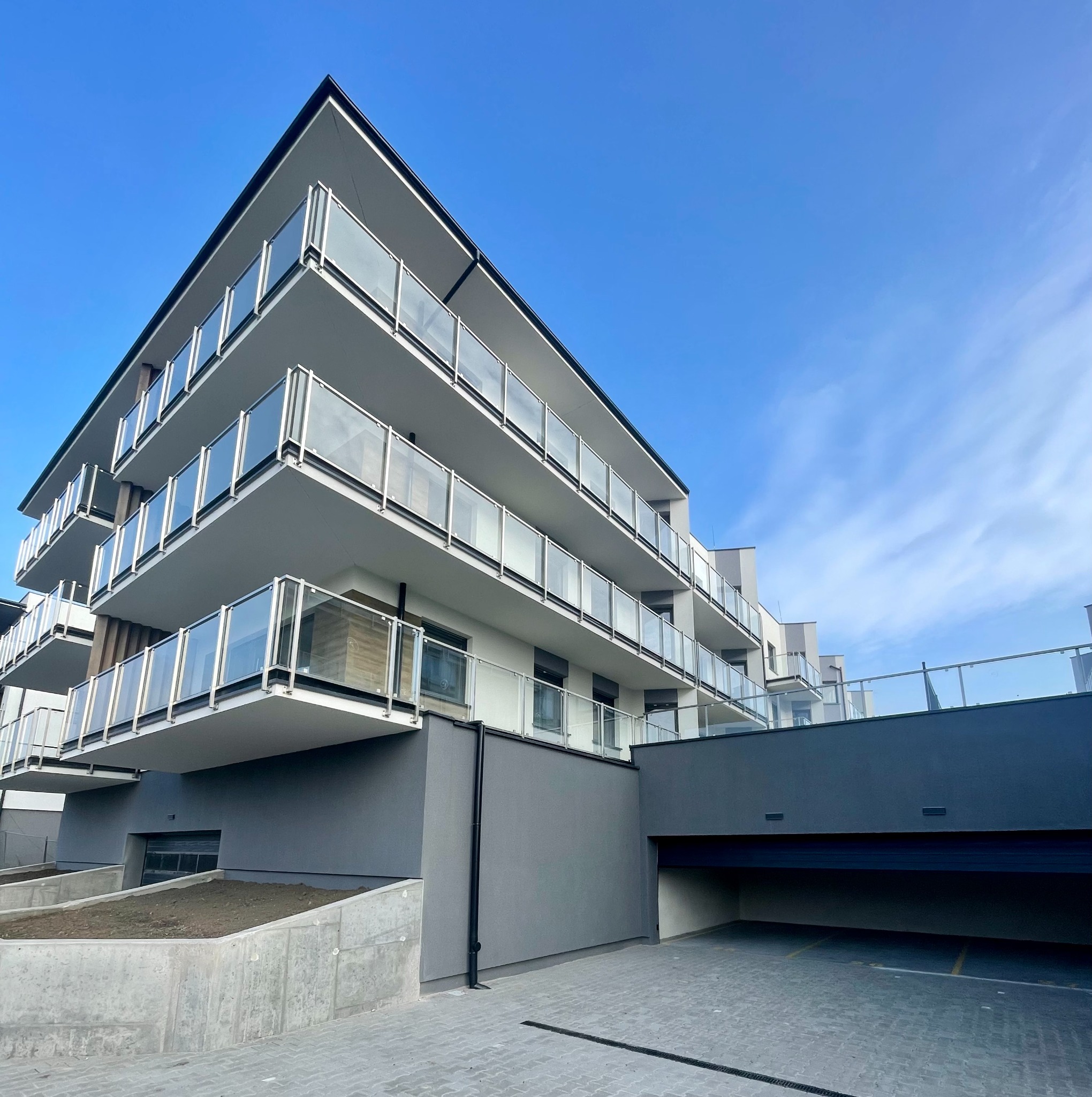 Nowoczesny budynek wielorodzinny z balkonami o szklanych balustradach i szarą elewacją, garaż podziemny, błękitne niebo w tle.
