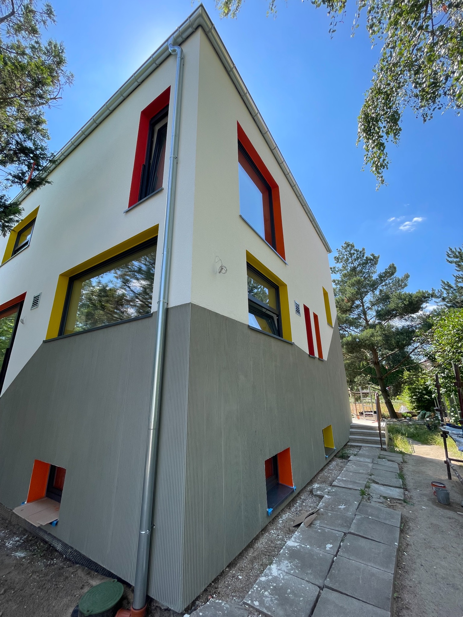 Nowoczesny dom z elewacją w odcieniach bieli i szarości, z kolorowymi obramowaniami okien (żółty, czerwony, pomarańczowy) i widocznym systemem rynnowym.