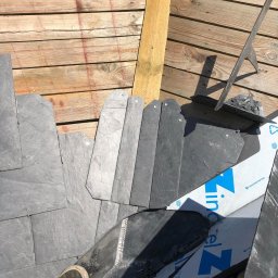 Hexagon Hub - Układanie łupka na dachu, widoczne drewniane łaty, narzędzie dekarskie i fragment buta robotnika.