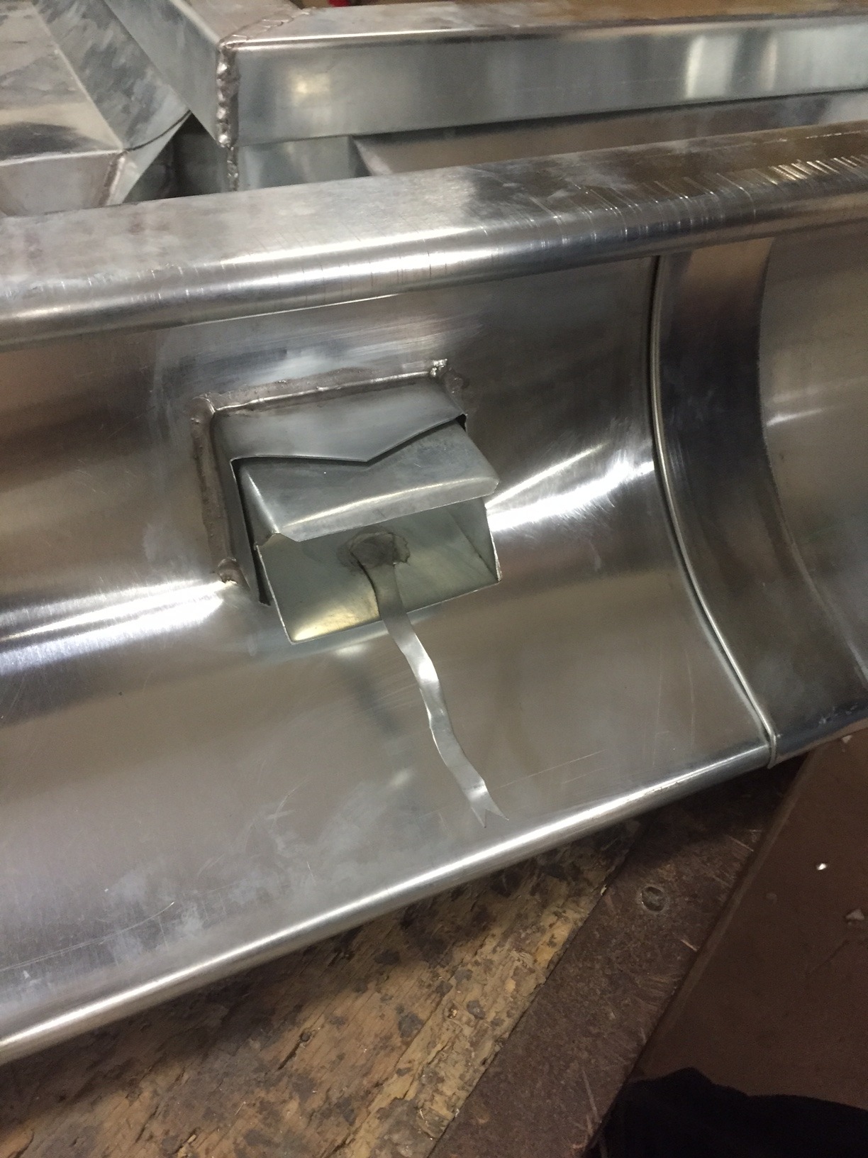 Szczegół metalowej rynny z zamontowanym odpływem, widoczne ślady spawania i elementy montażowe na tle drewnianego podłoża.