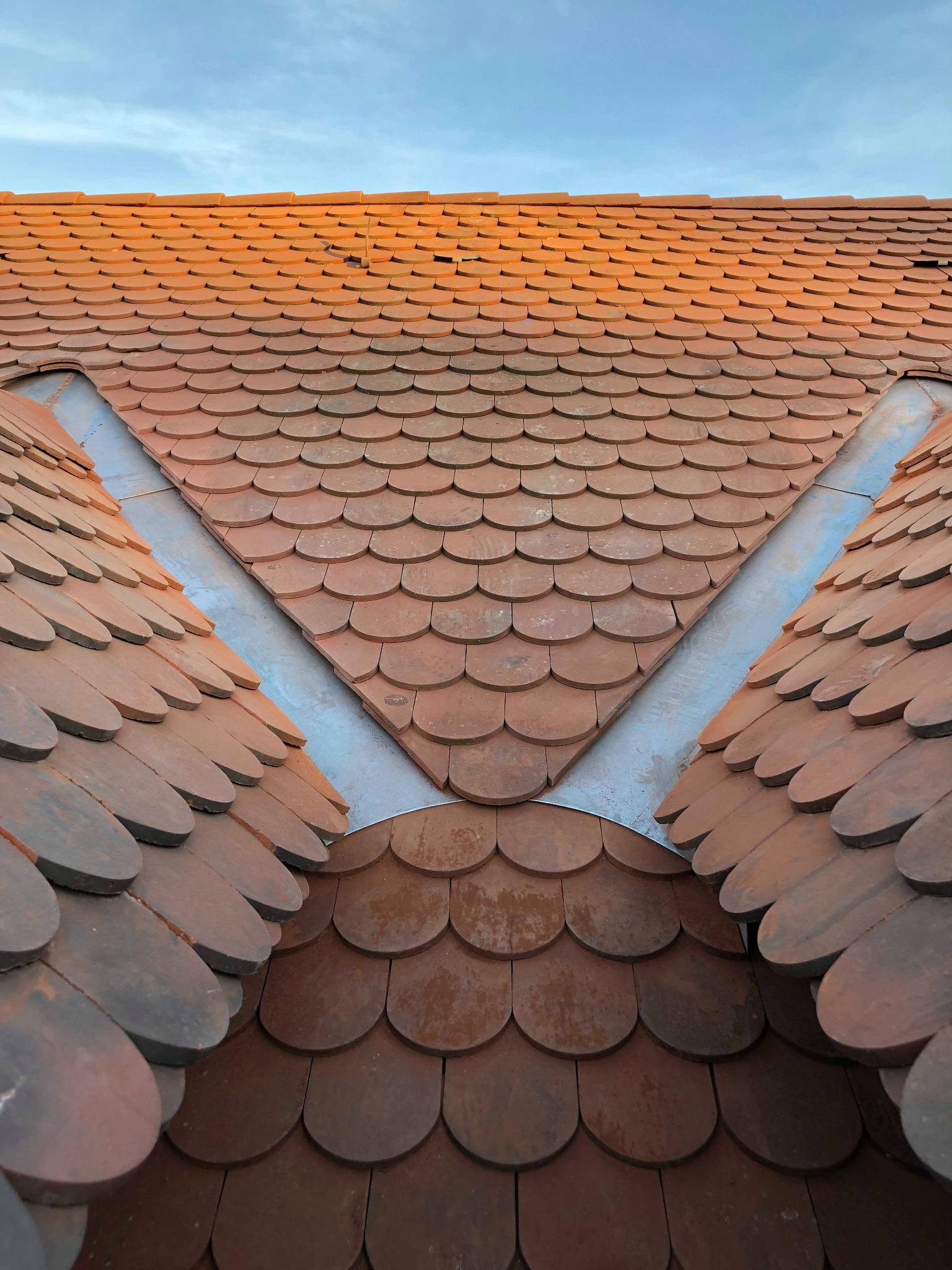 Widok z góry na brązowy dach pokryty dachówką karpiówką, z widocznym metalowym obramowaniem w kształcie litery V, na tle lekko zachmurzonego nieba.