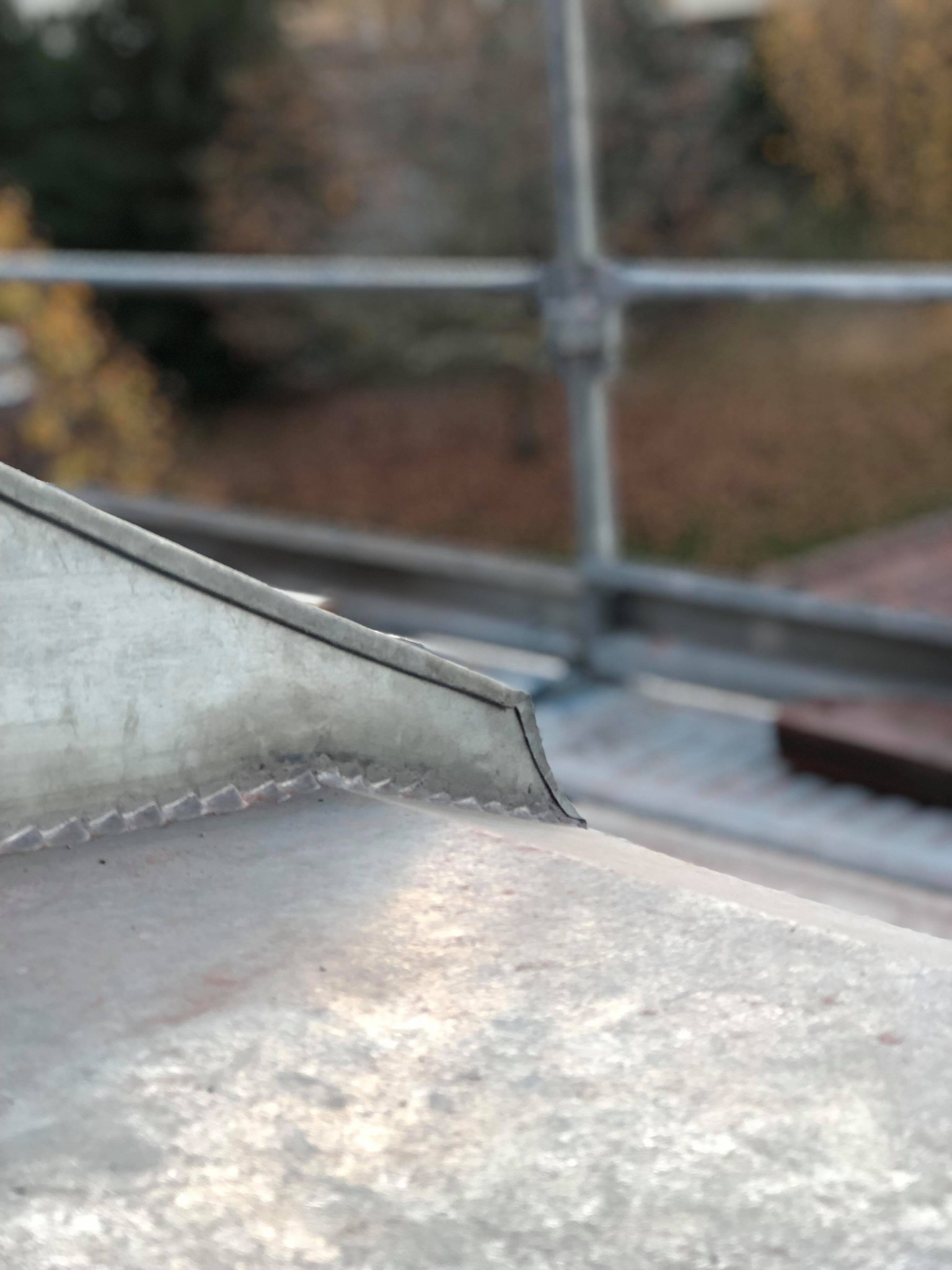 Szczegółowe zbliżenie na fragment metalowej obróbki blacharskiej dachu z widocznymi łączeniami, na tle rozmytego rusztowania i jesiennego krajobrazu.