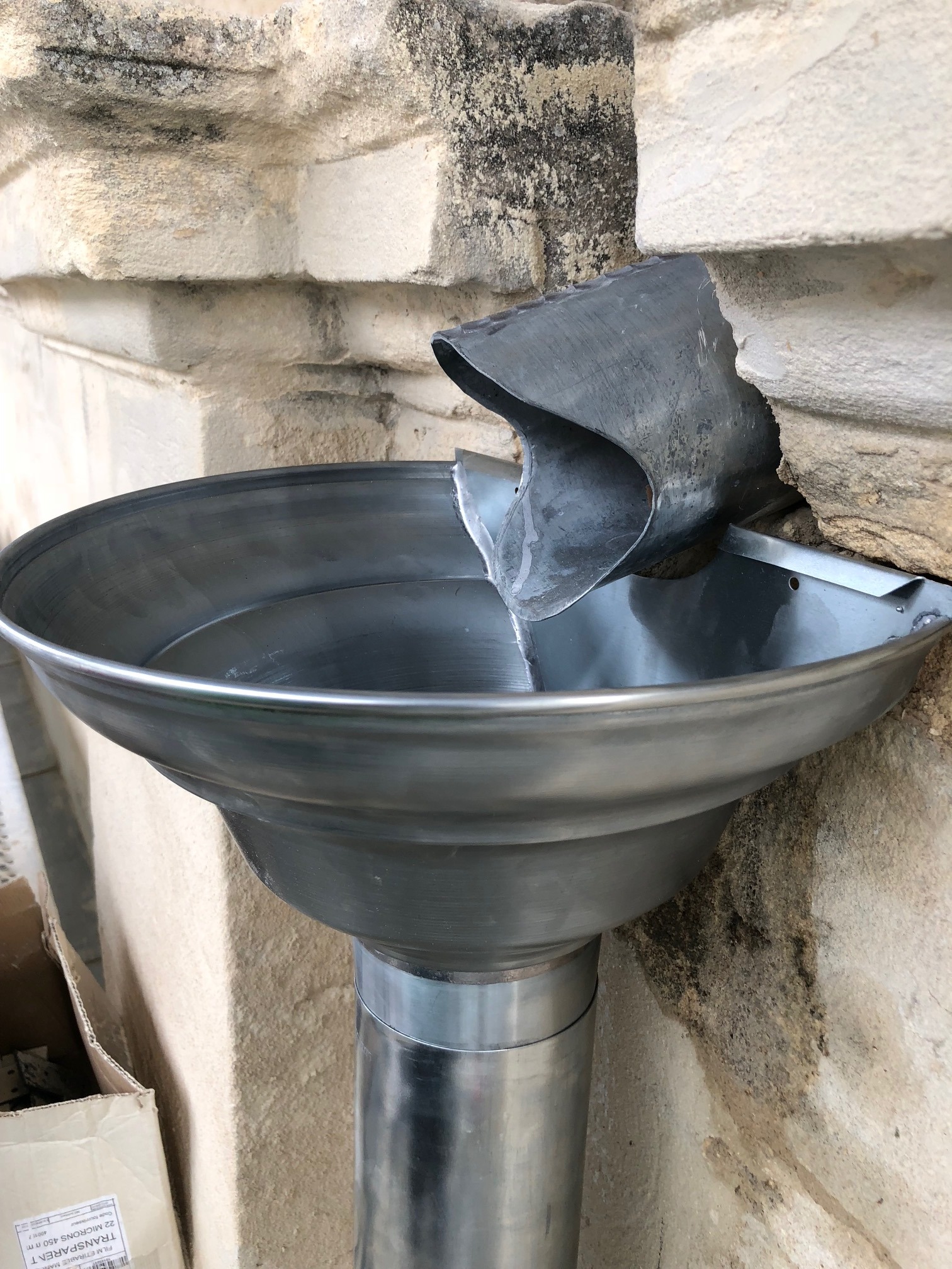 Metalowa rynna spustowa zamontowana na kamiennej ścianie, widoczne ślady po wodzie na ścianie i karton obok.