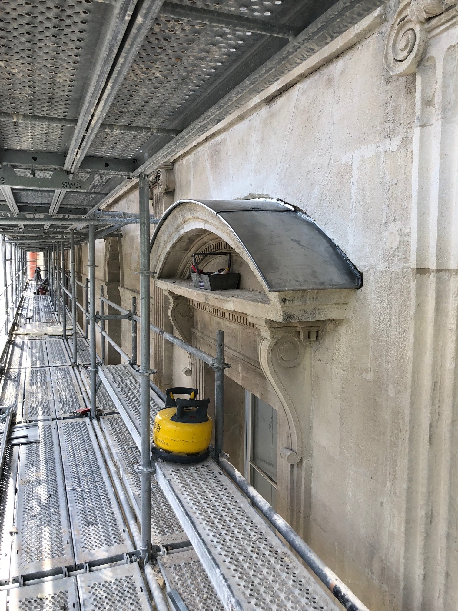 Renowacja kamiennej fasady budynku z elementami dekoracyjnymi, widoczna z poziomu rusztowania, z żółtą butlą gazową na platformie.