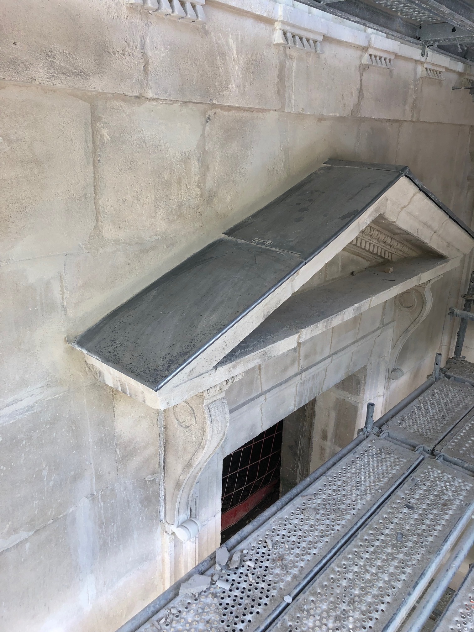 Kamienna fasada budynku w trakcie renowacji, z widocznym fragmentem dachu nad wejściem pokrytym blachą, rusztowanie wzdłuż ściany, detale architektoniczne.