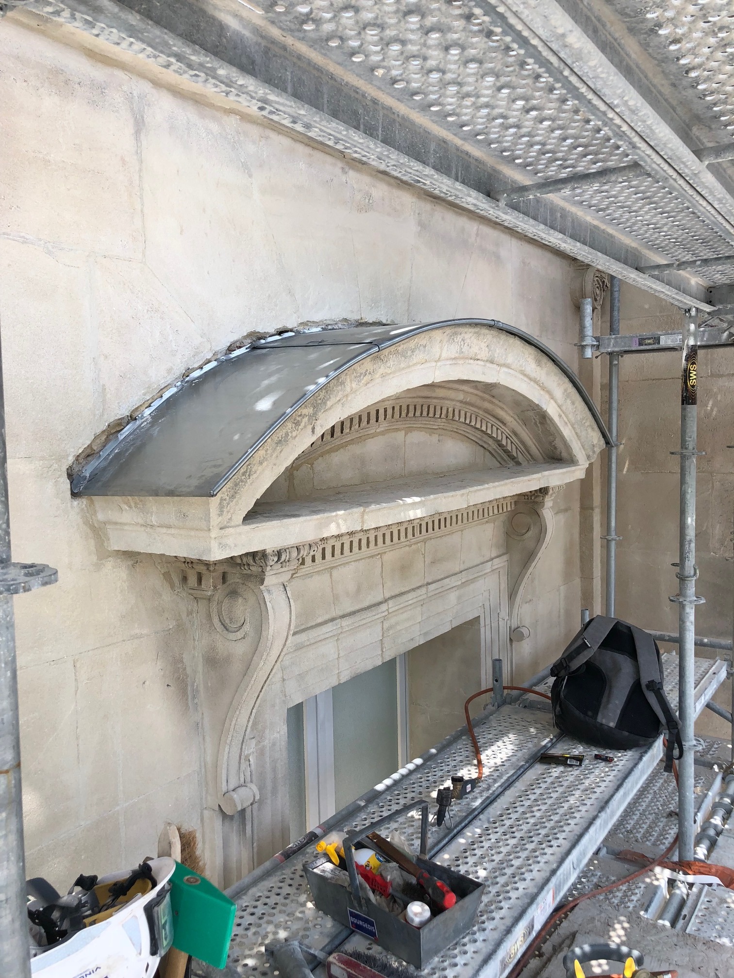 Odrestaurowany, ozdobny element architektoniczny nad oknem z nowym, metalowym zadaszeniem, widoczny ze rusztowania z narzędziami i plecakiem.