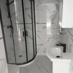 Nowoczesna łazienka z kabiną prysznicową z czarnymi profilami, umywalką na postumencie z czarną baterią i płytkami imitującymi marmur na ścianach i podłodze.