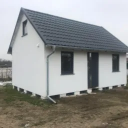 Nowo wybudowany, ocieplony dom jednorodzinny z szarą dachówką, widoczny w pochmurny dzień. Budynek postawiony na podwyższeniu z widoczną izolacją.