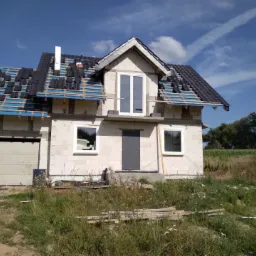 Niedokończony dom z białego bloczka z częściowo ułożonym, czarnym dachem, widoczne niebieskie membrany dachowe i biały komin, okna plastikowe białe, szara brama garażowa.