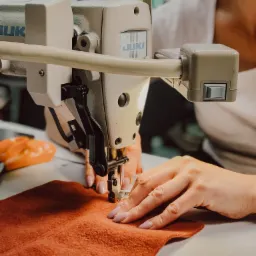 Praca na maszynie do szycia JUKI: zbliżenie na szycie brązowego materiału, dłonie z jasnoróżowym manicure, widoczny fragment białej koszulki osoby szyjącej.