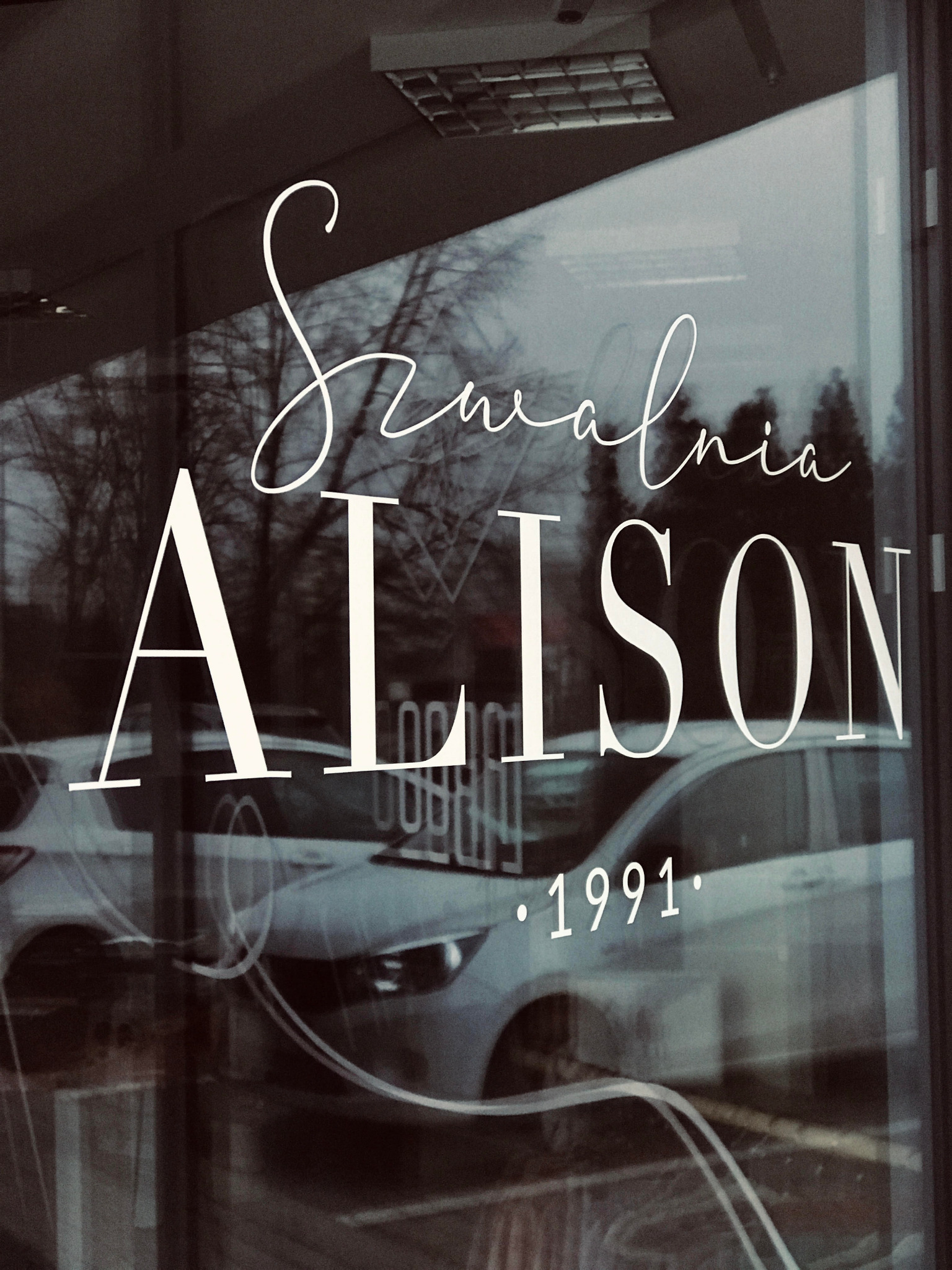 Szwalnia Alison z rokiem założenia 1991, naklejona na szklanej witrynie z odbiciem samochodów i drzew.