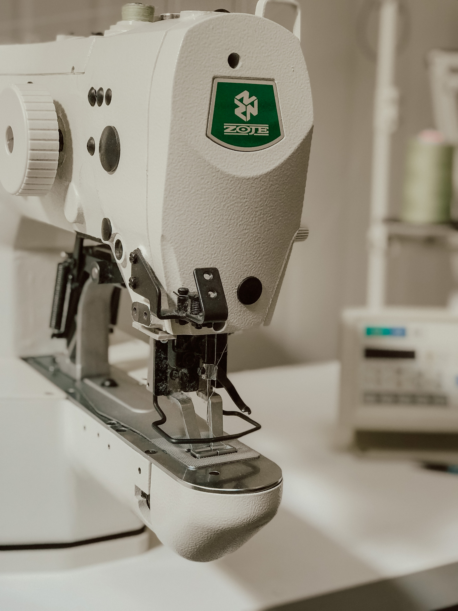 Zbliżenie na białą maszynę do szycia marki ZOJE z widoczną igłą i mechanizmem, z rolką nici i inną maszyną w tle.