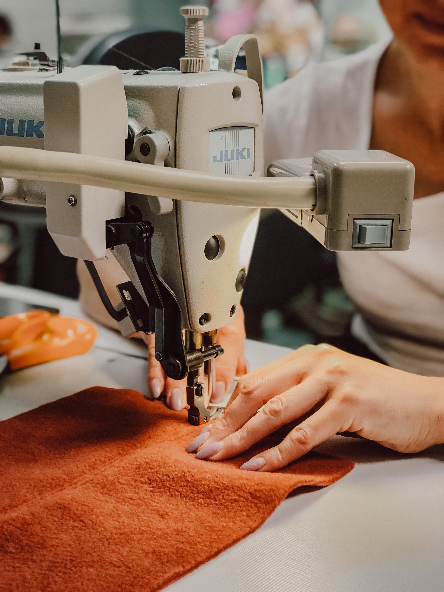 Praca na maszynie do szycia JUKI: zbliżenie na szycie brązowego materiału, dłonie z jasnoróżowym manicure, widoczny fragment białej koszulki osoby szyjącej.
