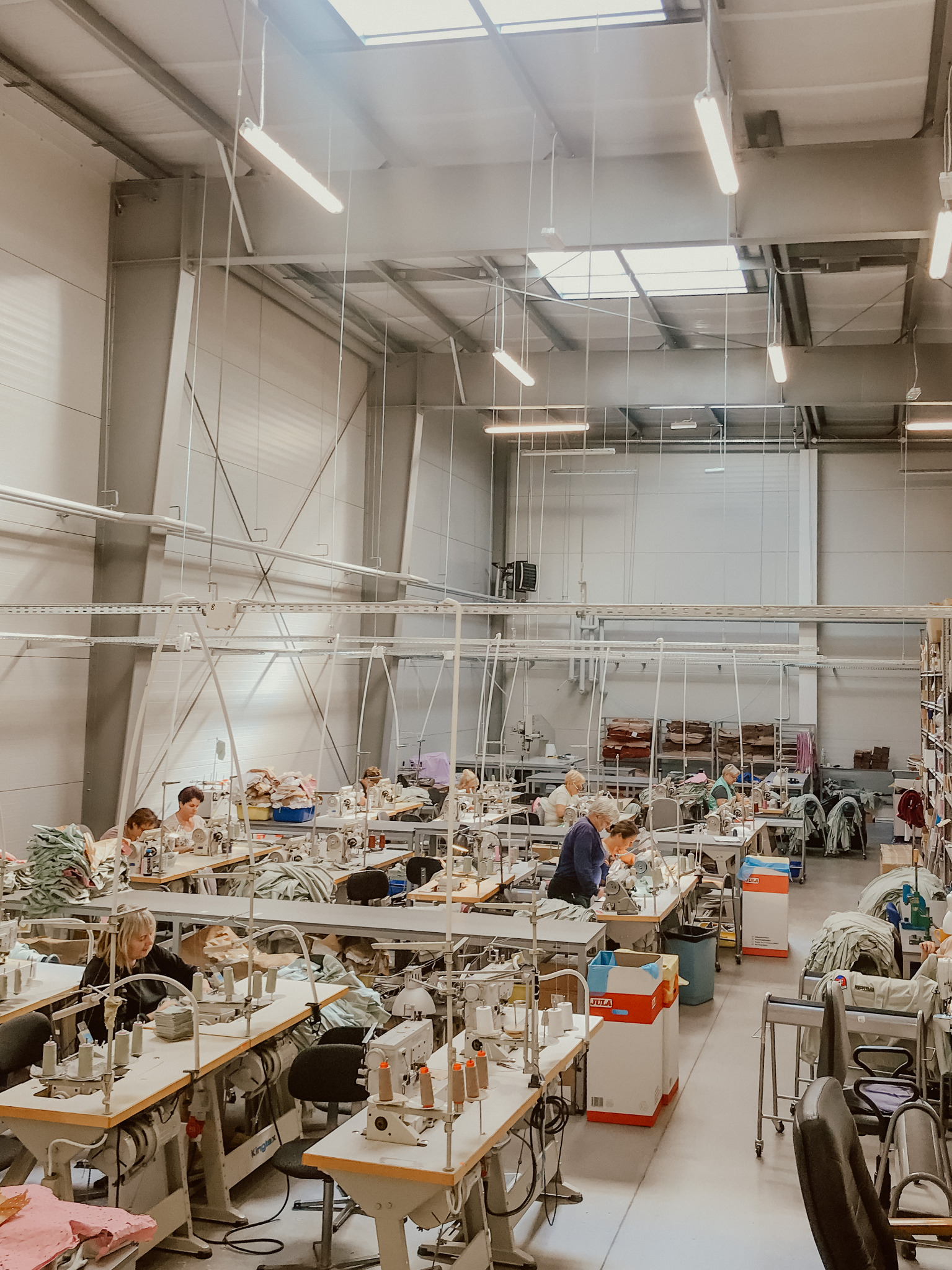 Przestronne wnętrze hali produkcyjnej z rzędami stanowisk pracy wyposażonych w maszyny do szycia, pracownicy przy pracy, oświetlenie jarzeniowe i okna dachowe.