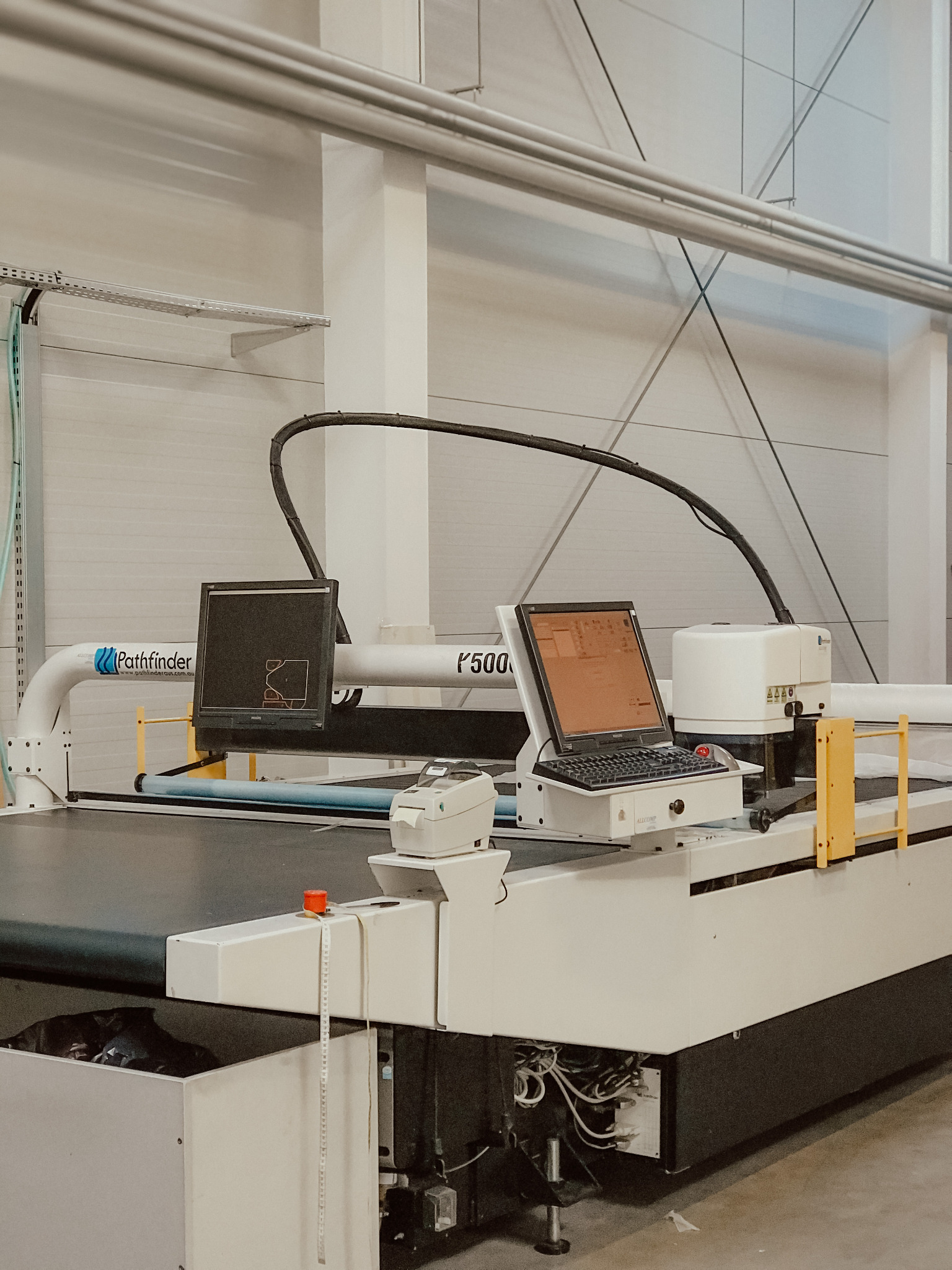 Duża, biała maszyna do cięcia tkanin Pathfinder F500 z komputerem, monitorem, drukarką etykiet i miarką, widoczna w hali produkcyjnej.