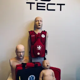 Manekiny do nauki pierwszej pomocy: dorosły w kamizelce Act+Fast, dorosły bez kamizelki i niemowlę, obok torba ratownicza z symbolem gwiazdy życia, na ścianie logo firmy Protect.