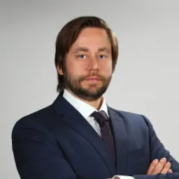 Portret mężczyzny w ciemnym garniturze i fioletowym krawacie, z brodą i włosami zaczesanymi do tyłu, pozującego na jasnoszarym tle ze skrzyżowanymi rękami.