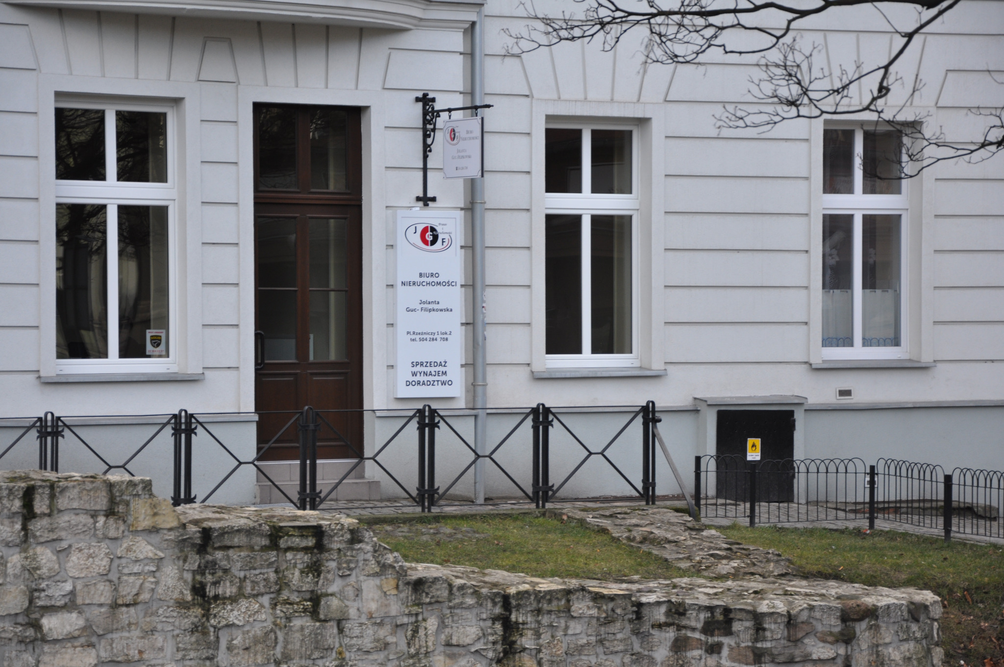 Elewacja budynku biura nieruchomości z szyldem informacyjnym oferującym sprzedaż, wynajem i doradztwo, widoczna kamienna barierka.