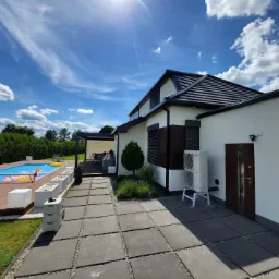 Biała pompa ciepła Panasonic Aquarea na metalowym stelażu, zainstalowana przy ścianie nowoczesnego domu z ciemną stolarką okienną i drzwiami wejściowymi, na zewnątrz kostka brukowa, w tle basen...