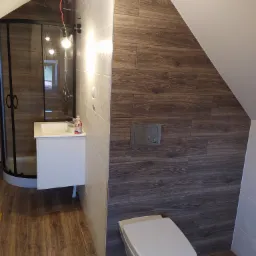Wykończona łazienka na poddaszu z drewnopodobnymi płytkami, kabina prysznicowa z czarnymi ramami, biała umywalka z szafką, wisząca toaleta i niedokończone oświetlenie sufitowe