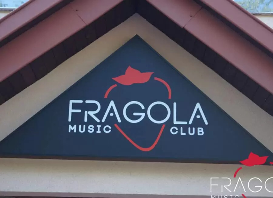 Trójkątny szyld z nazwą 'FRAGOLA MUSIC CLUB' w kolorze białym i czerwonym, umieszczony nad wejściem do lokalu.