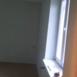 Świeżo pomalowane wnętrze pokoju z drewnianą podłogą i oknem, widoczne ślady taśmy malarskiej na ścianie i para narzędzi na parapecie.