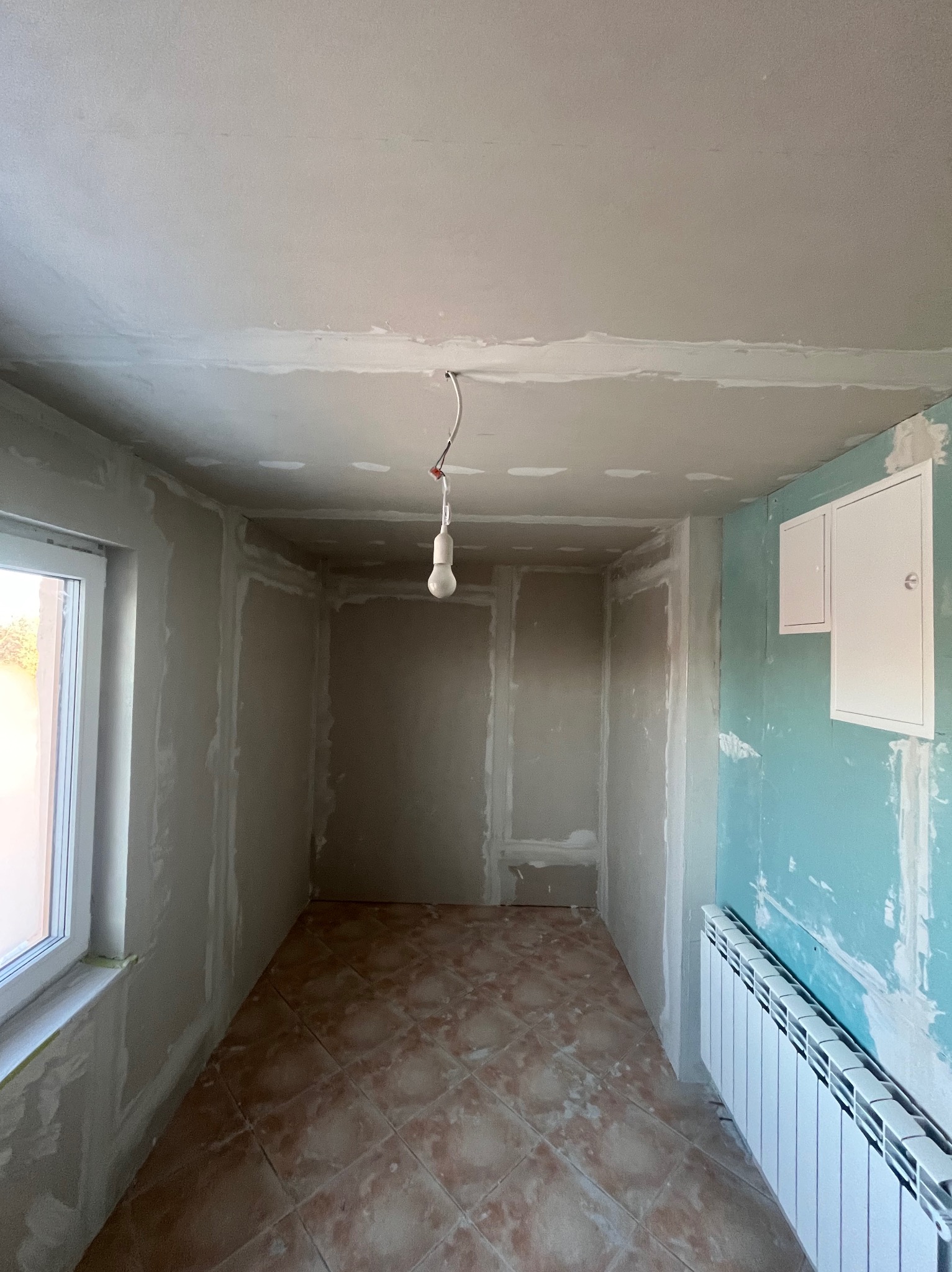 Pokój w trakcie prac przygotowawczych: ściany po szpachlowaniu, widoczne łączenia płyt gipsowych, grzejnik, okno i wisząca żarówka. Podłoga wyłożona płytkami.