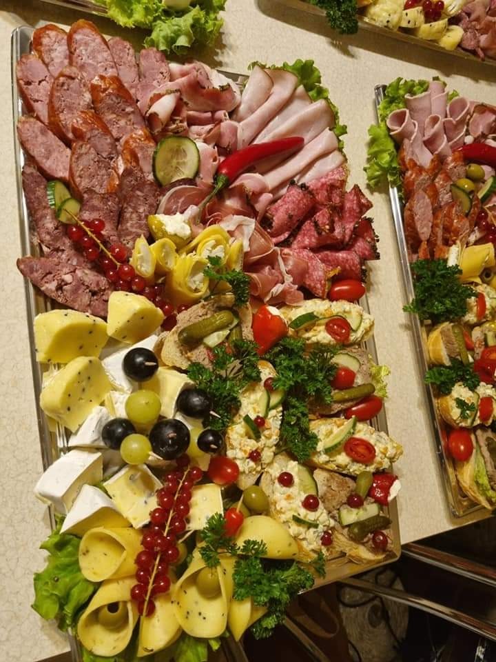 Dwa srebrne półmiski z bogatym wyborem wędlin, serów, kanapek i owoców, udekorowane zieloną sałatą i czerwoną porzeczką. Na pierwszym planie półmisek z różnymi rodzajami kiełbas, szynki, serów...