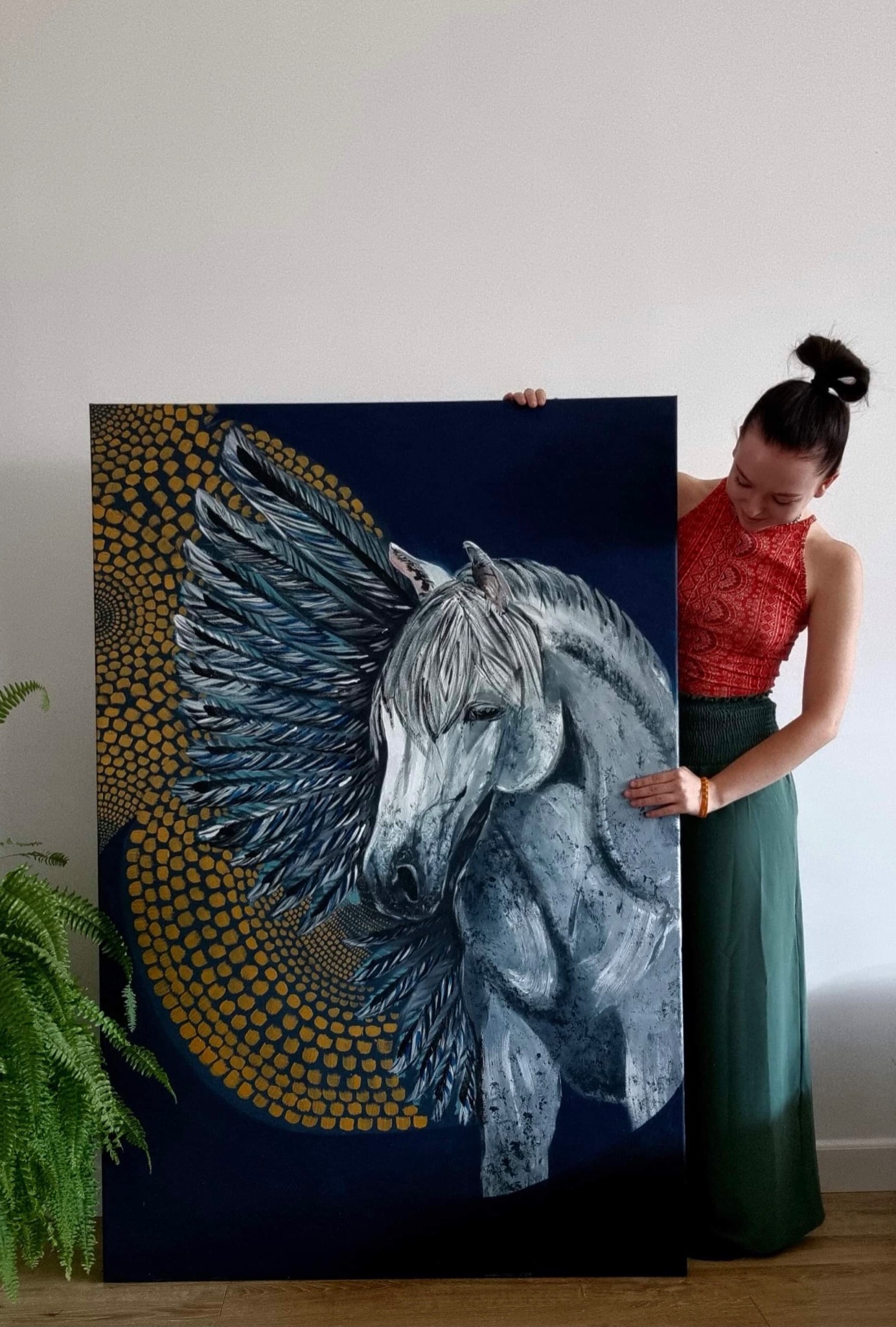 Młoda kobieta prezentuje obraz olejny przedstawiający skrzydlatego konia na ciemnoniebieskim tle z geometrycznym wzorem w odcieniach złota, stojąc obok zielonej rośliny doniczkowej.