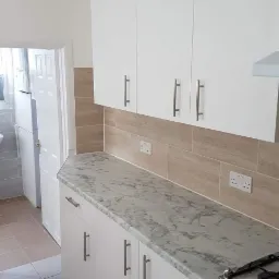 Wykończona kuchnia z białymi szafkami, marmurowym blatem i beżowymi płytkami na ścianie, widok na łazienkę z otwartymi drzwiami.