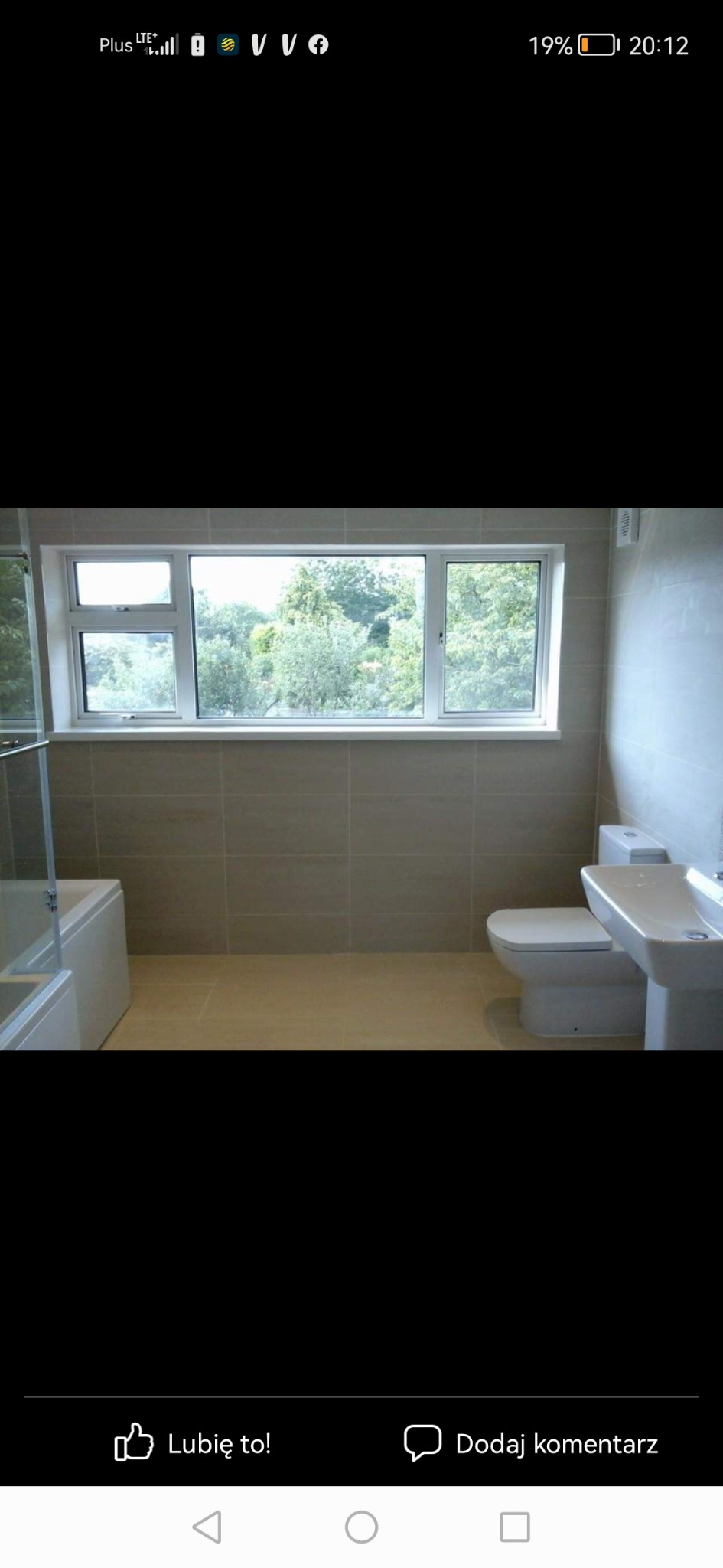 Jasna łazienka z beżowymi płytkami, widok na zieleń za oknem, biała umywalka i toaleta, częściowo widoczna kabina prysznicowa.