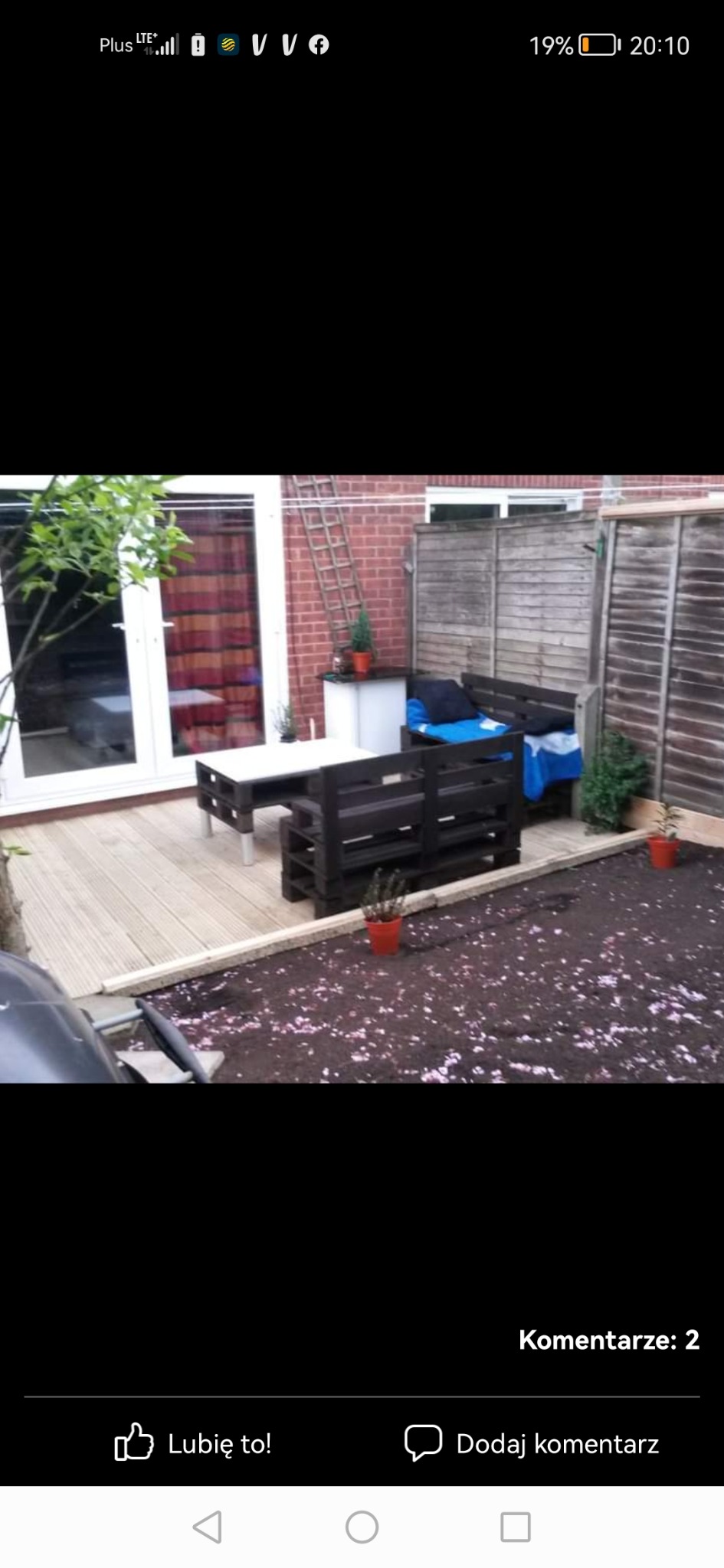 Drewniany taras z meblami ogrodowymi wykonanymi z palet, stolik z metalowymi nogami, ziemia z opadłymi płatkami kwiatów, widoczne drzwi balkonowe, drabina oparta o ścianę budynku.