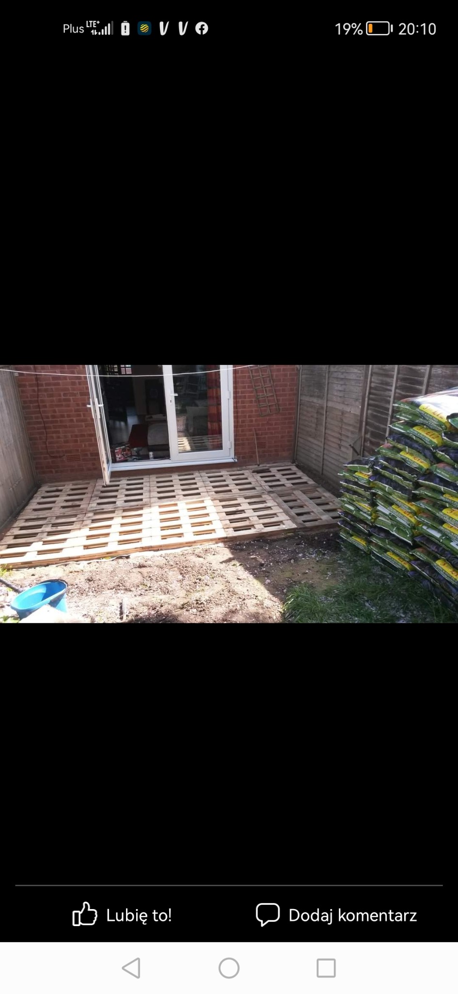 Tymczasowy taras z palet przed wejściem do domu z cegły, obok stos worków z ziemią i drewniany płot.