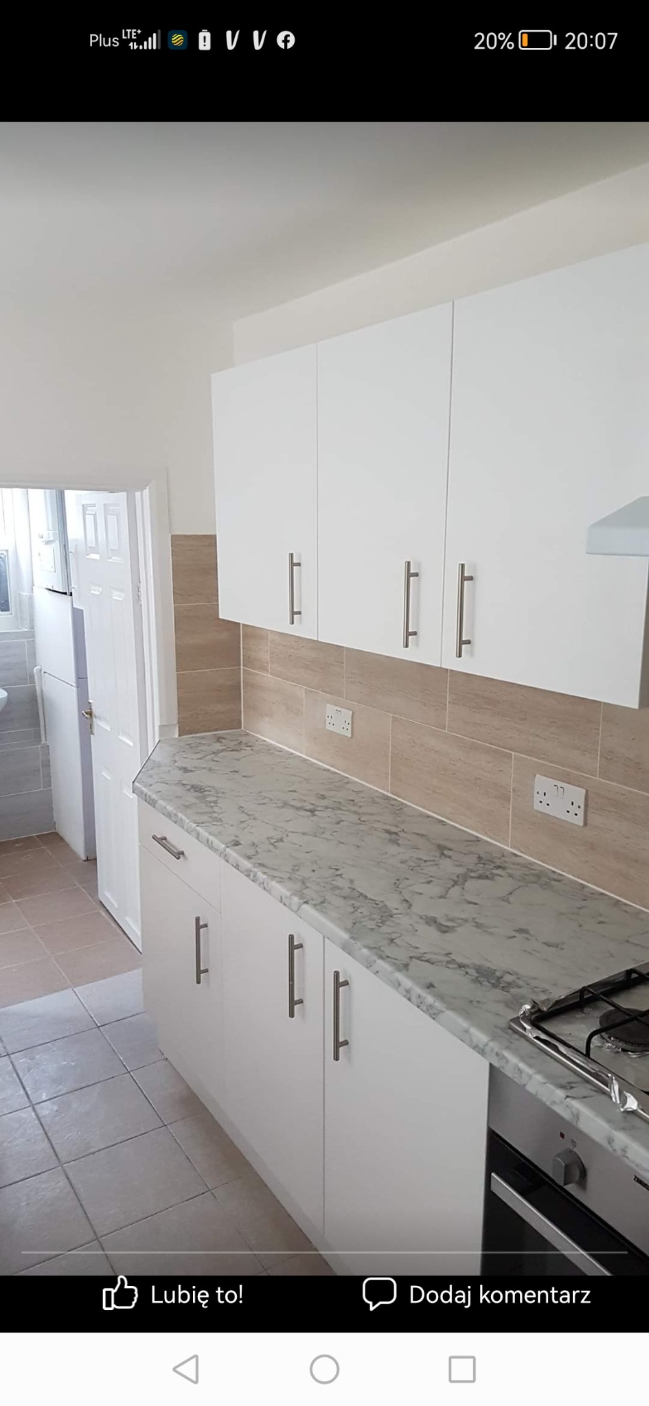 Wykończona kuchnia z białymi szafkami, marmurowym blatem i beżowymi płytkami na ścianie, widok na łazienkę z otwartymi drzwiami.