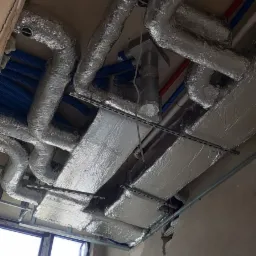 Instalacja wentylacji i rekuperacji pod sufitem podwieszanym, widoczne rury izolowane folią aluminiową, niebieskie rury wentylacyjne karbowane oraz czerwone rury, surowy stan wykończenia wnętrza.