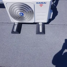 Biała jednostka zewnętrzna klimatyzatora marki Kaisai, zamontowana na dachu na gumowych podkładkach antywibracyjnych, z widocznym cieniem fotografa.