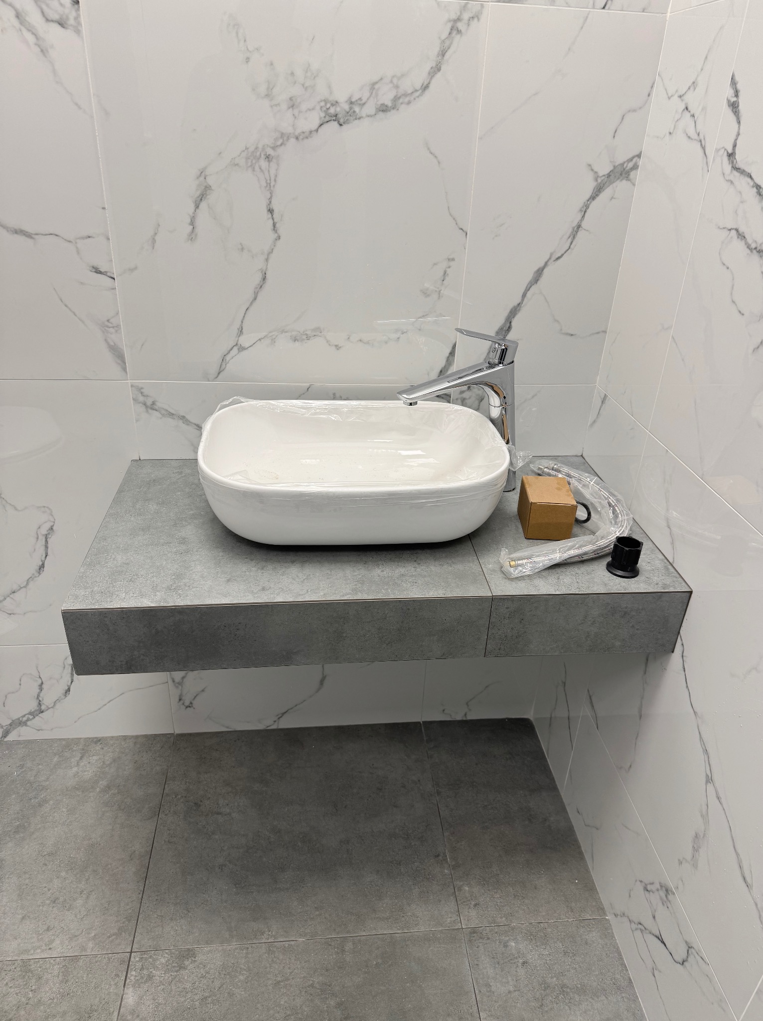 Nowoczesna łazienka z białą umywalką nablatową owiniętą folią ochronną, chromowaną baterią i akcesoriami montażowymi na szarym blacie z płytek, marmurowe płytki na ścianach i szara podłoga z płytek.