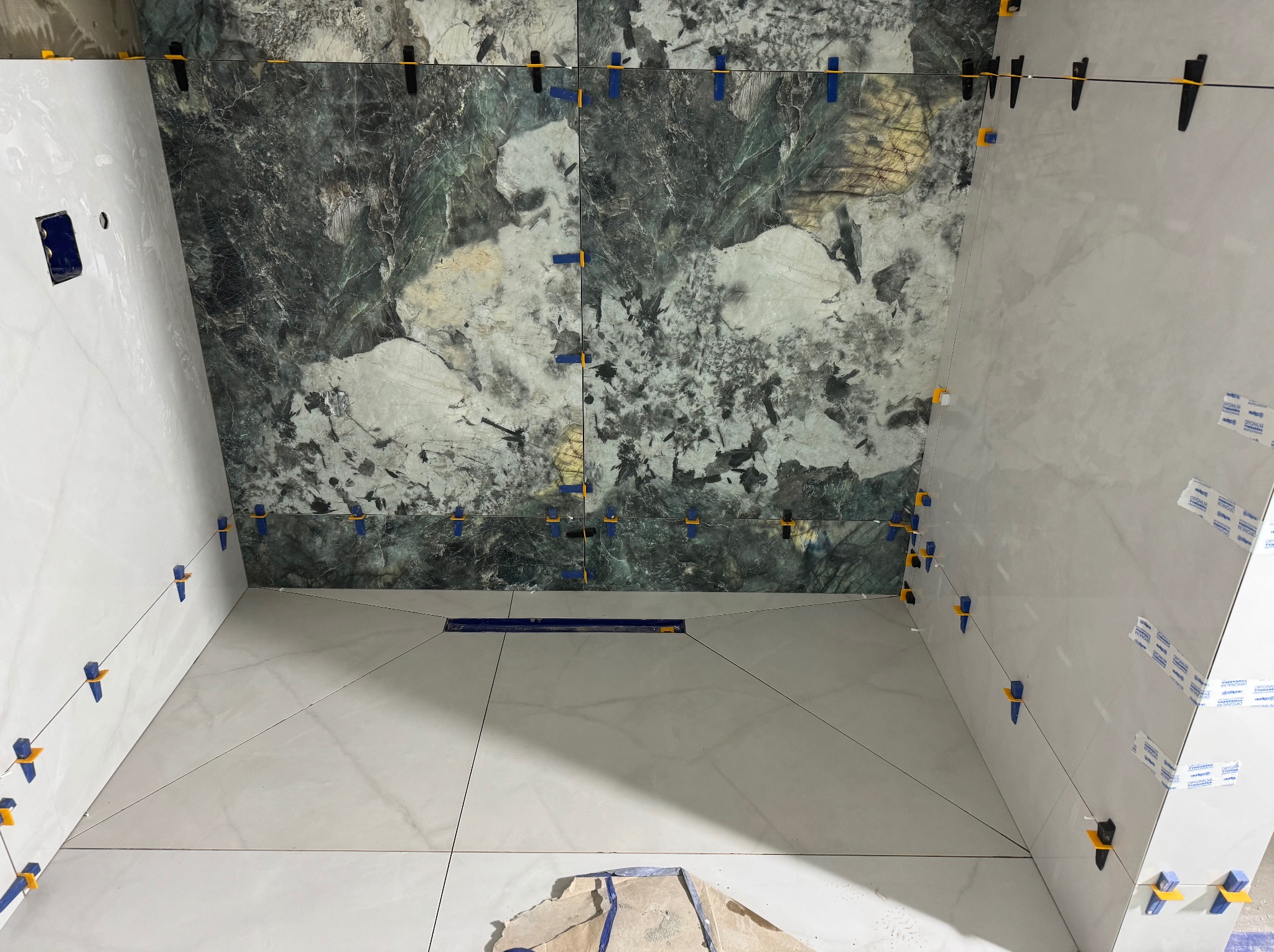 Montaż płytek w łazience: białe płytki na ścianach i podłodze, zielono-szare płytki imitujące kamień na jednej ze ścian, widoczne systemy poziomowania płytek.