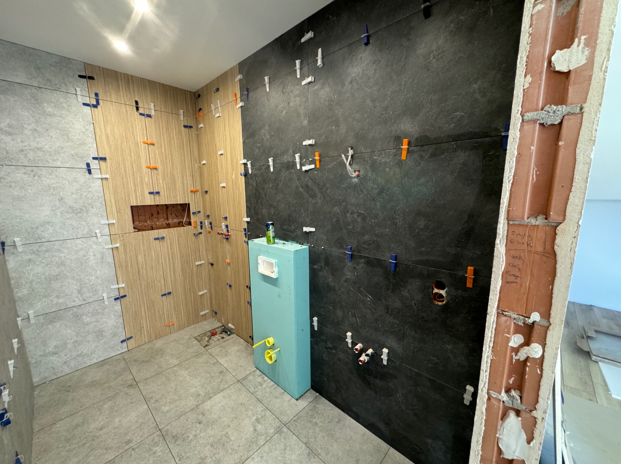 Łazienka w trakcie kładzenia płytek: ściany częściowo wyłożone płytkami imitującymi drewno, szarym kamieniem i ciemnym gresem, widoczne krzyżyki dystansowe, stelaż podtynkowy WC w kolorze turkusowym.