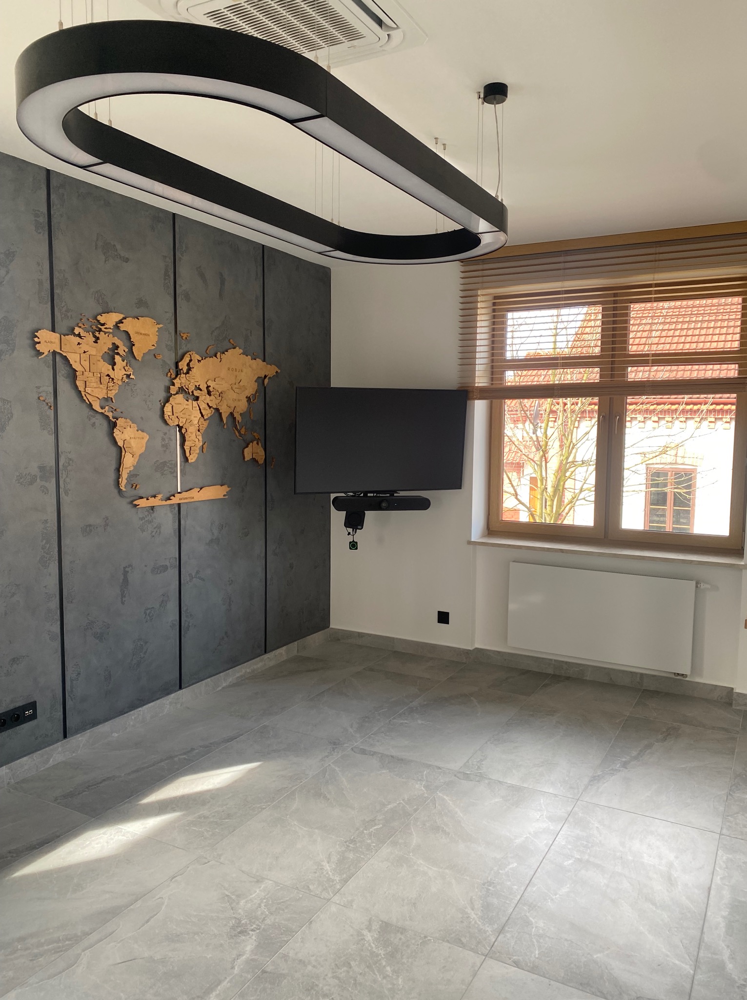 Pokój po remoncie z szarą ścianą z drewnianą mapą świata, telewizorem na ścianie, oknem z drewnianymi żaluzjami i nowoczesną lampą wiszącą.
