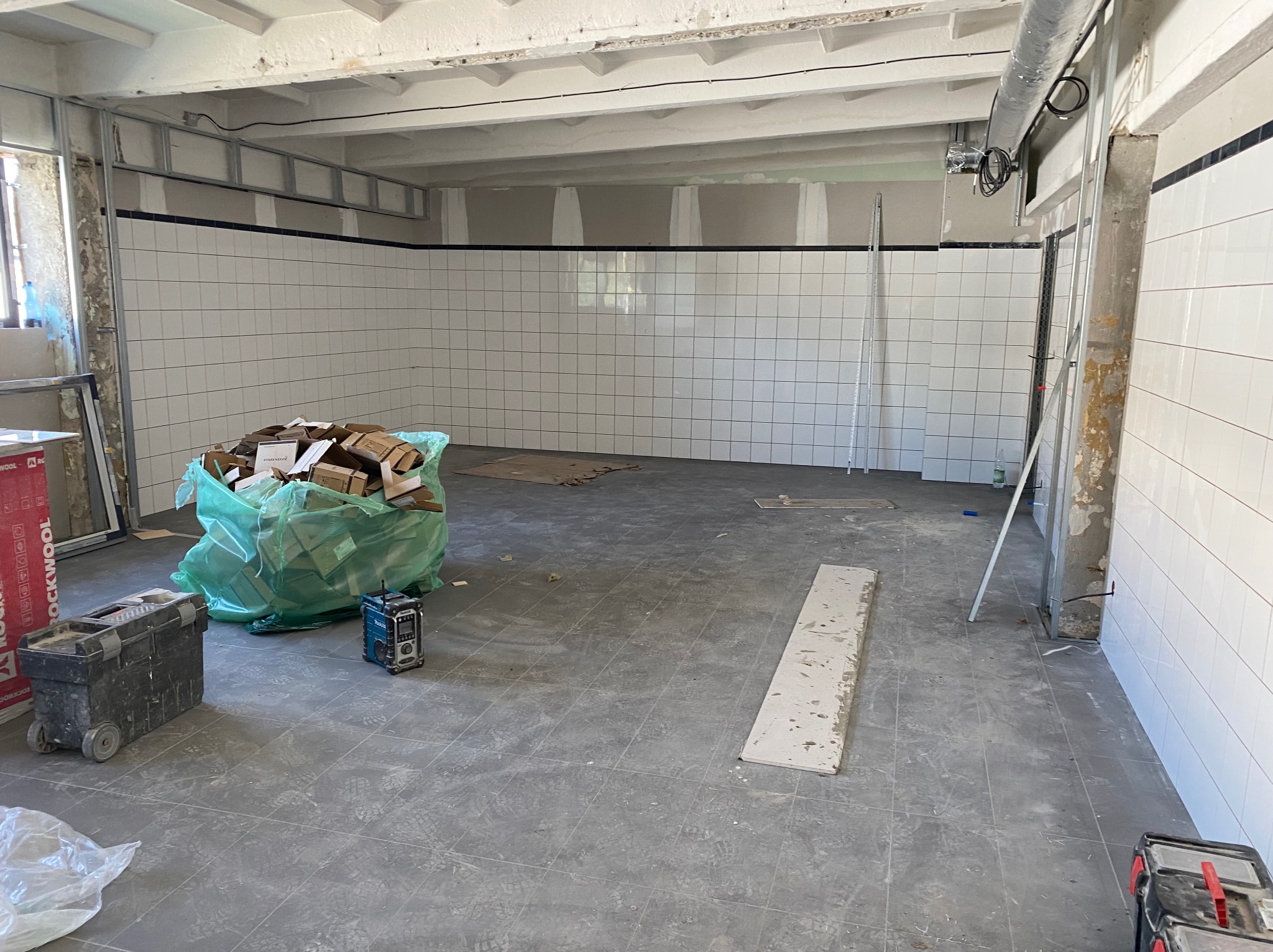 Wnętrze w trakcie prac wykończeniowych, ściany wyłożone białymi płytkami, widoczny worek z odpadami, skrzynka narzędziowa i radio budowlane.