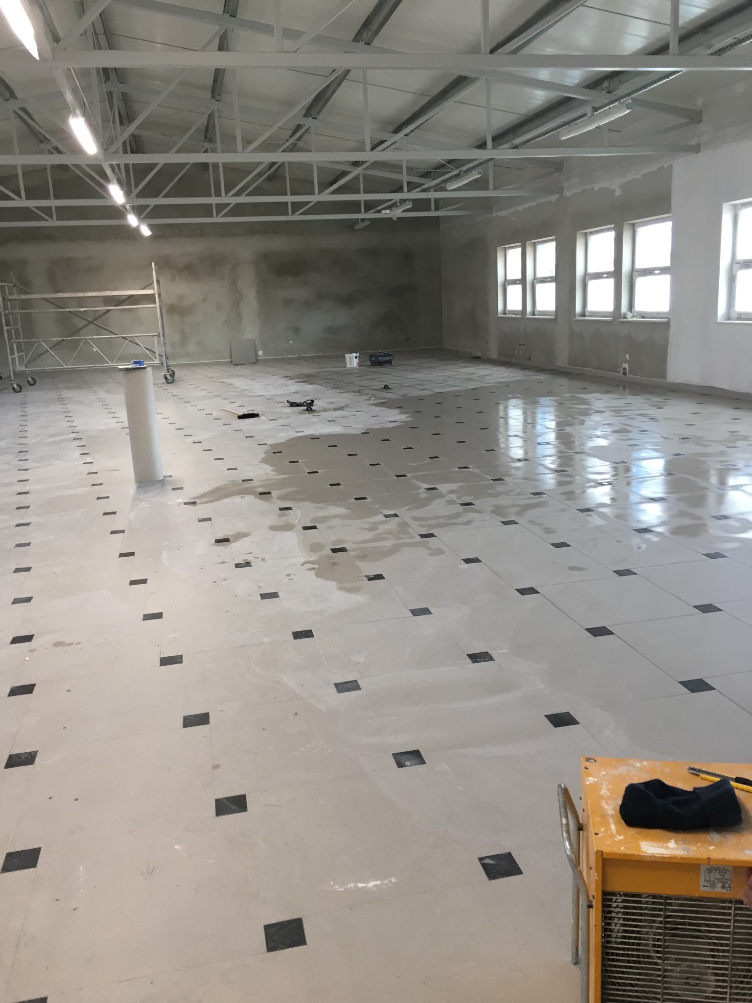Wnętrze dużego pomieszczenia w trakcie prac wykończeniowych z ułożoną jasną podłogą z ciemnymi, kwadratowymi wstawkami, mokra powierzchnia, widoczne rusztowanie, grzejnik budowlany i narzędzia.